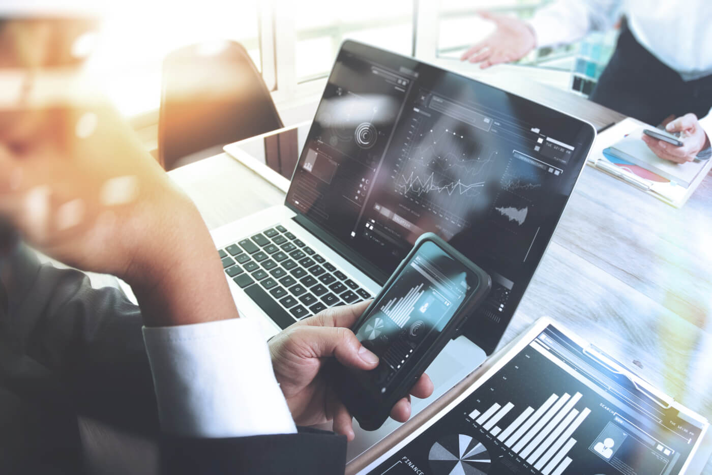 person sitting in front of computer holding cell phone with graphs and data on both screens
